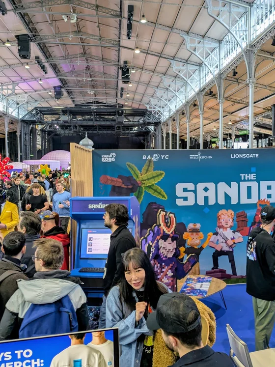 The Sandbox stand at NFT Paris, crowded with visitors, the arcade cabinet in use in front of a large pixel-art backdrop.